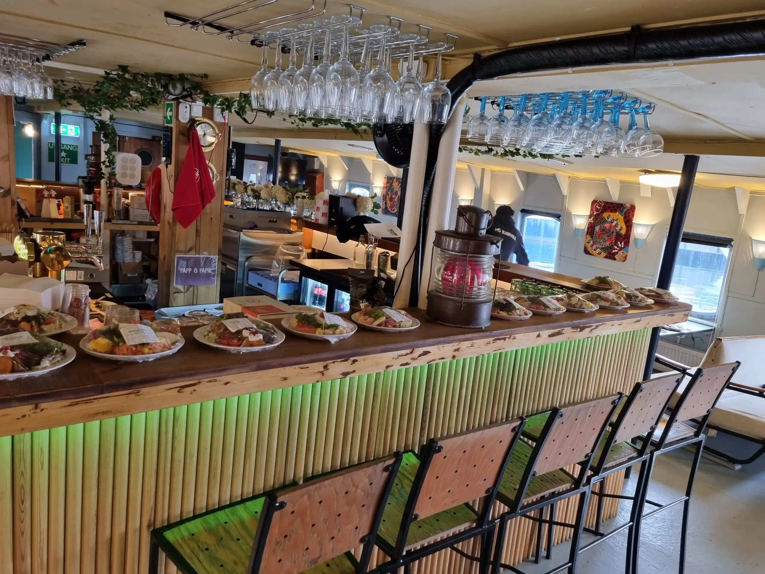 En bar med trefront dekorert med grønne lys, hvor tallerkener med ulike retter står klare, og glass henger i stativ over bardisken.